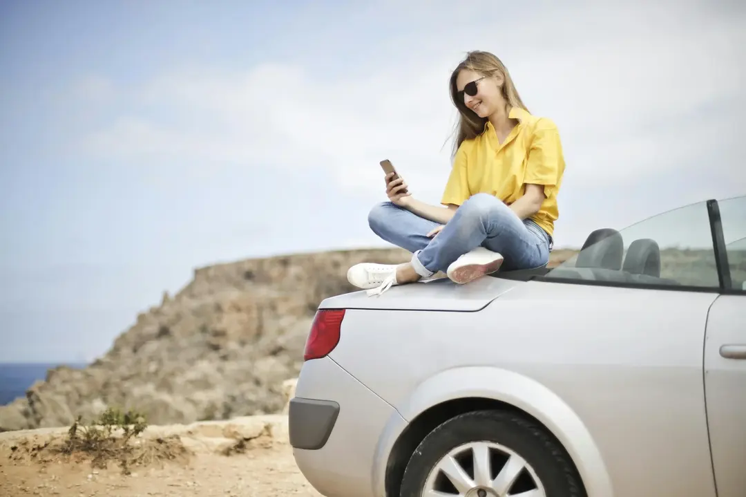 Mulher jovem, vestindo uma blusa amarela e jeans, sentada no porta-malas de um conversível prateado enquanto segura um celular. Ao fundo, um cenário natural com penhascos sob um céu claro.