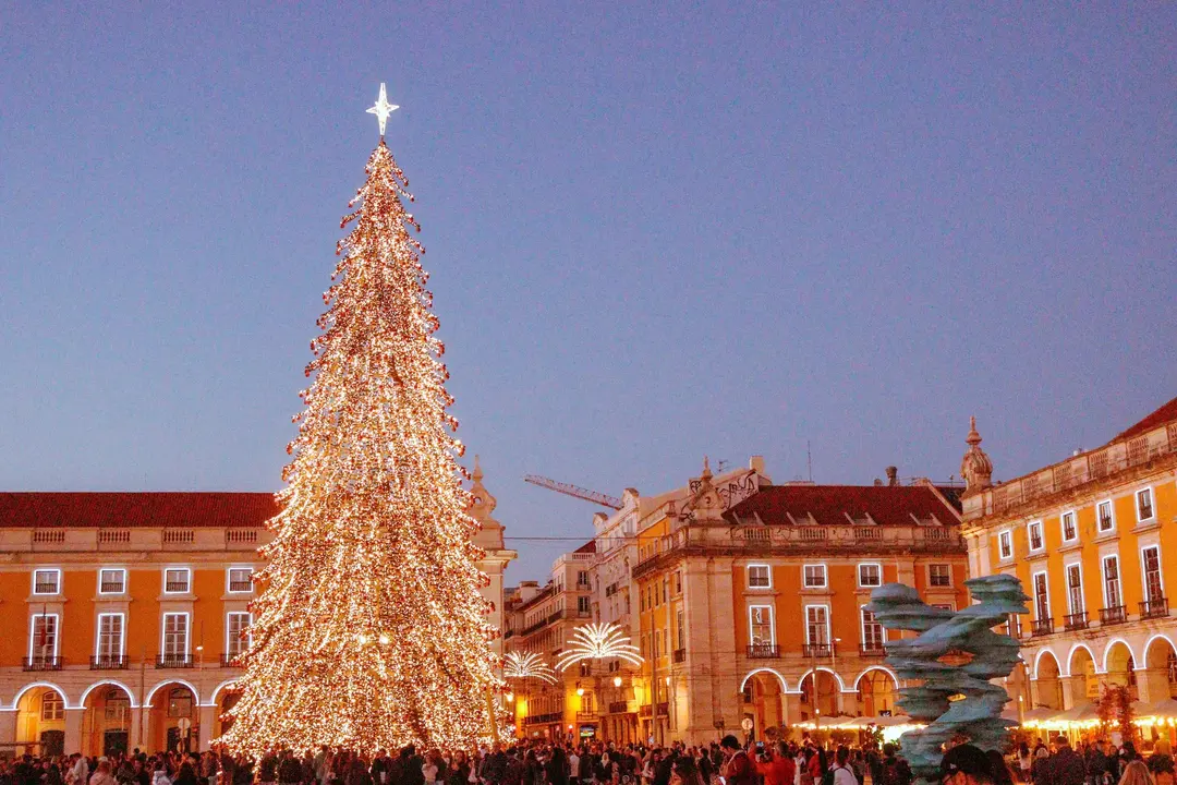 Uma árvore de Natal iluminada no centro da Praça do Comércio em Lisboa, com edifícios históricos ao fundo e um céu limpo no início da noite.
