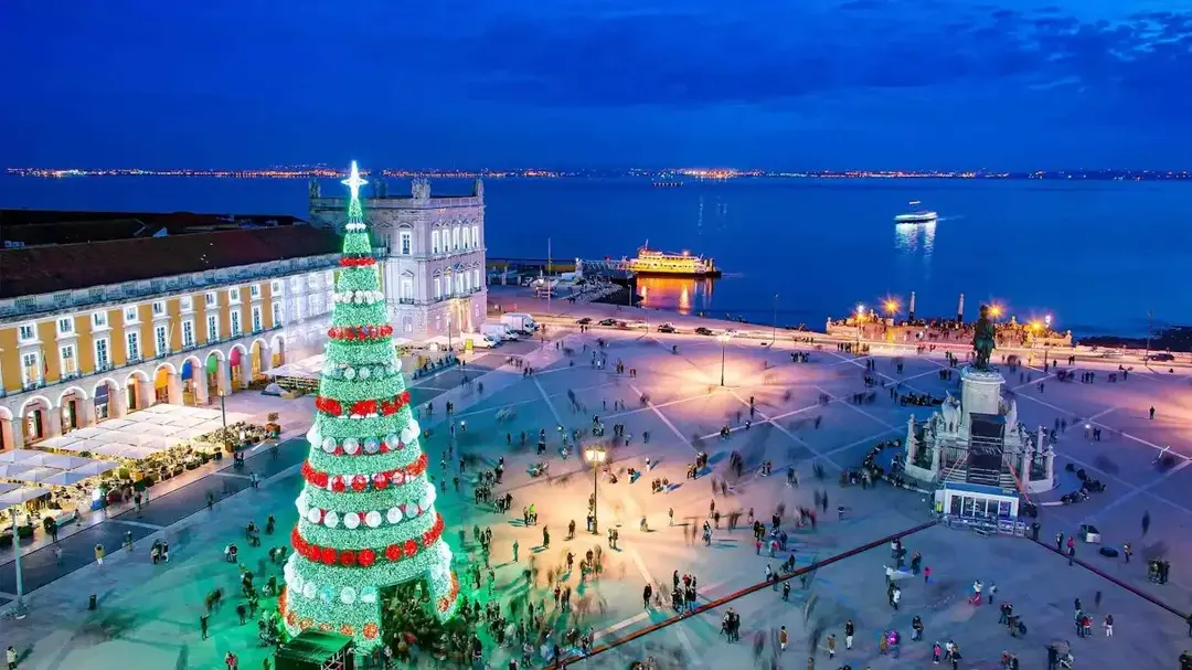 Uma árvore de Natal gigante e iluminada no centro da Praça do Comércio, com o rio Tejo ao fundo, em uma noite clara.