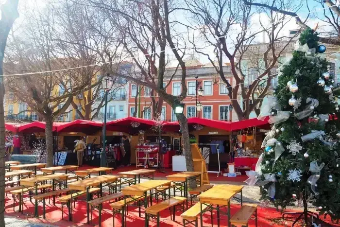 Um mercado natalino com barraquinhas decoradas e uma árvore de Natal ao fundo, em uma praça ao ar livre.