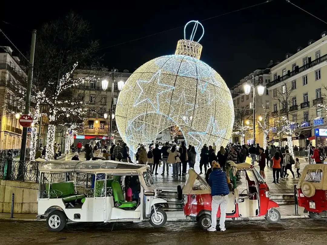 Uma grande instalação luminosa em formato de bola de Natal decorada com luzes brancas e douradas, cercada por tuk-tuks e pedestres em uma praça movimentada à noite.
