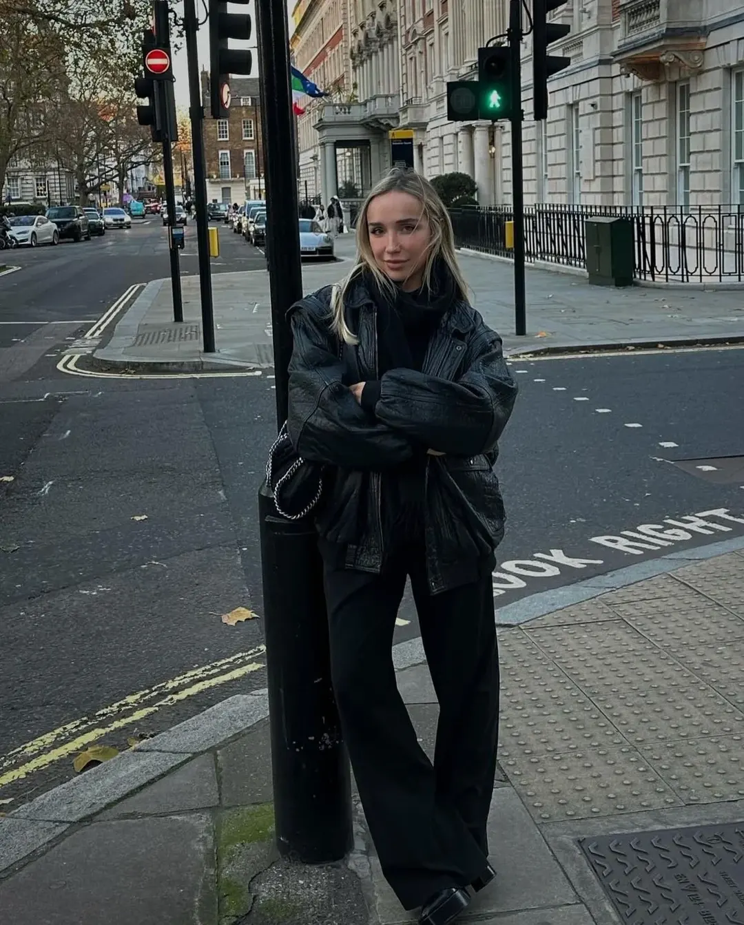 Mulher de braços cruzados em um poste, vestida com calça e casaco pretos