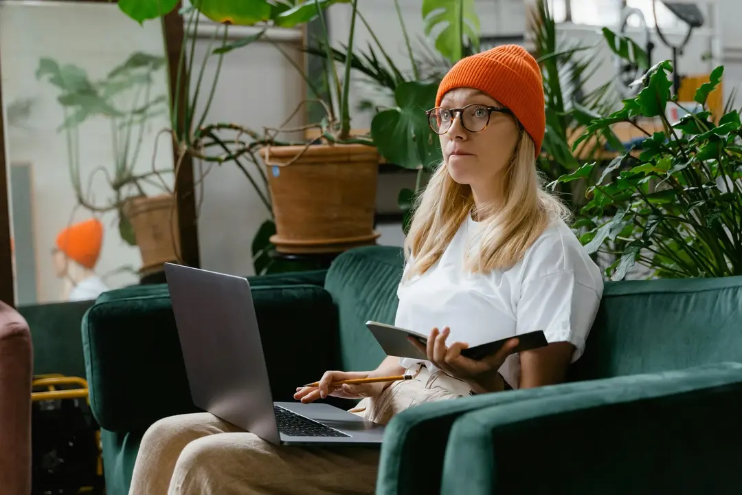 Mulher com gorro laranja sentada em um ambiente aconchegante, explorando destinos no laptop enquanto faz anotações.