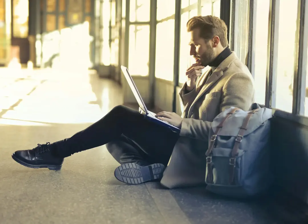 Homem sentado no chão do aeroporto, focado em seu laptop, planejando detalhes de viagem.