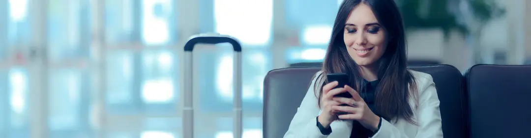 Mulher sorridente usando o celular enquanto está na espera em um aeroporto.