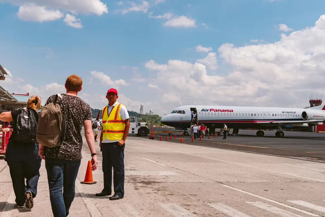 Passageiros caminhando na pista em direção ao avião, com céu azul ao fundo e um funcionário do aeroporto em destaque.